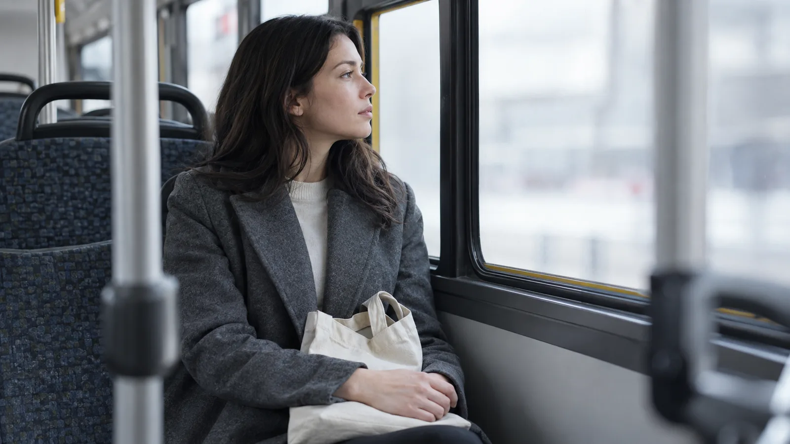 Woman in a grey wool coat sitting by a bus window with a canvas tote on her lap, gazing outside.
