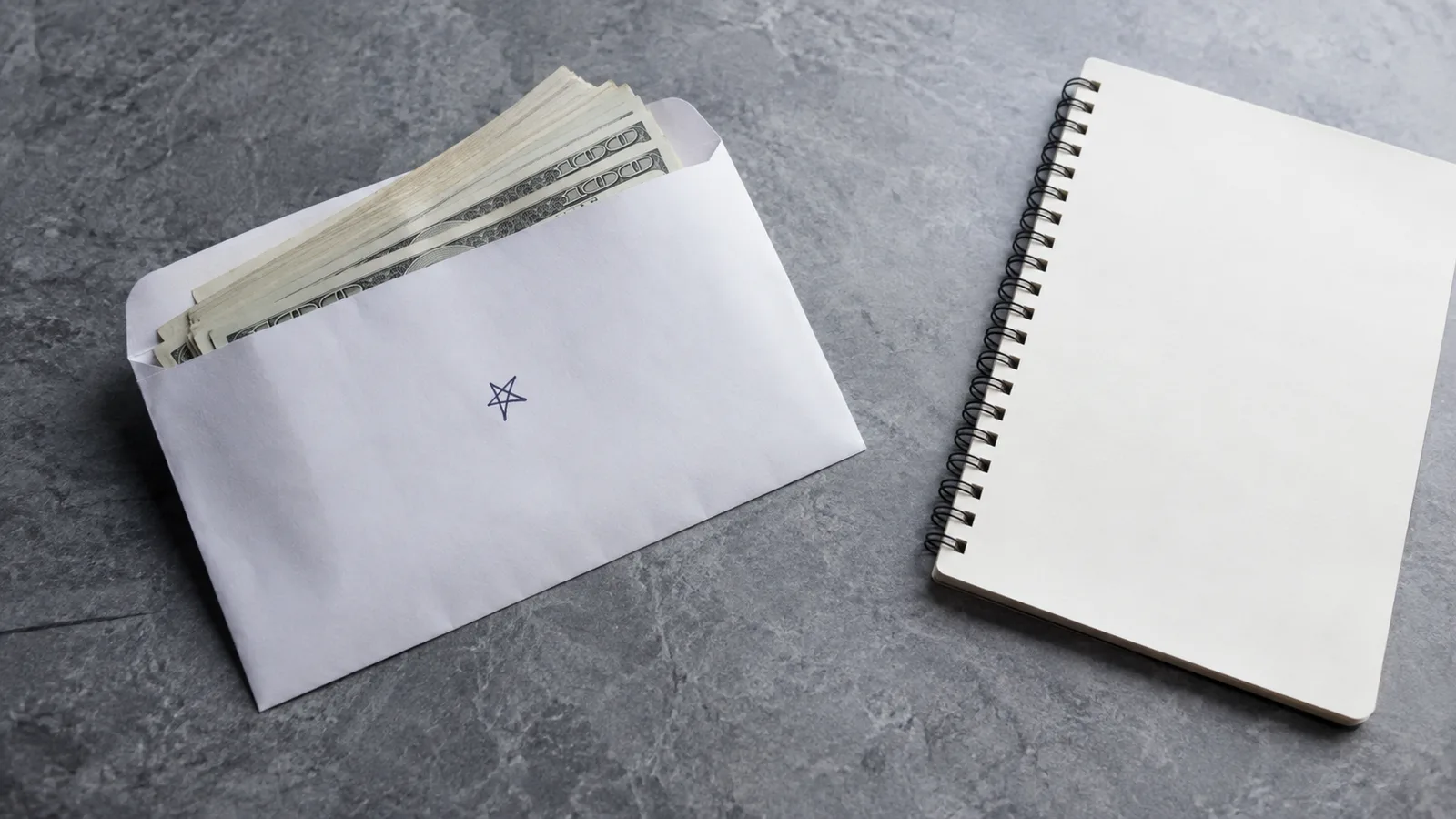 White envelope stuffed with banknotes lying next to a blank spiral notebook on a grey stone surface.